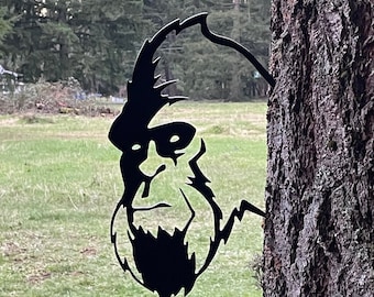 May include: Black metal silhouette of a Bigfoot or Sasquatch head with a beard, attached to a tree trunk.