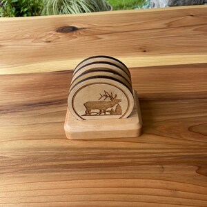 May include: Set of four wooden coasters with a laser-etched design of an elk standing in a forest. The coasters are stacked on a wooden stand.
