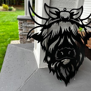May include: Black metal silhouette of a Highland cow head with a bow on its head. The silhouette is mounted on a gray surface.