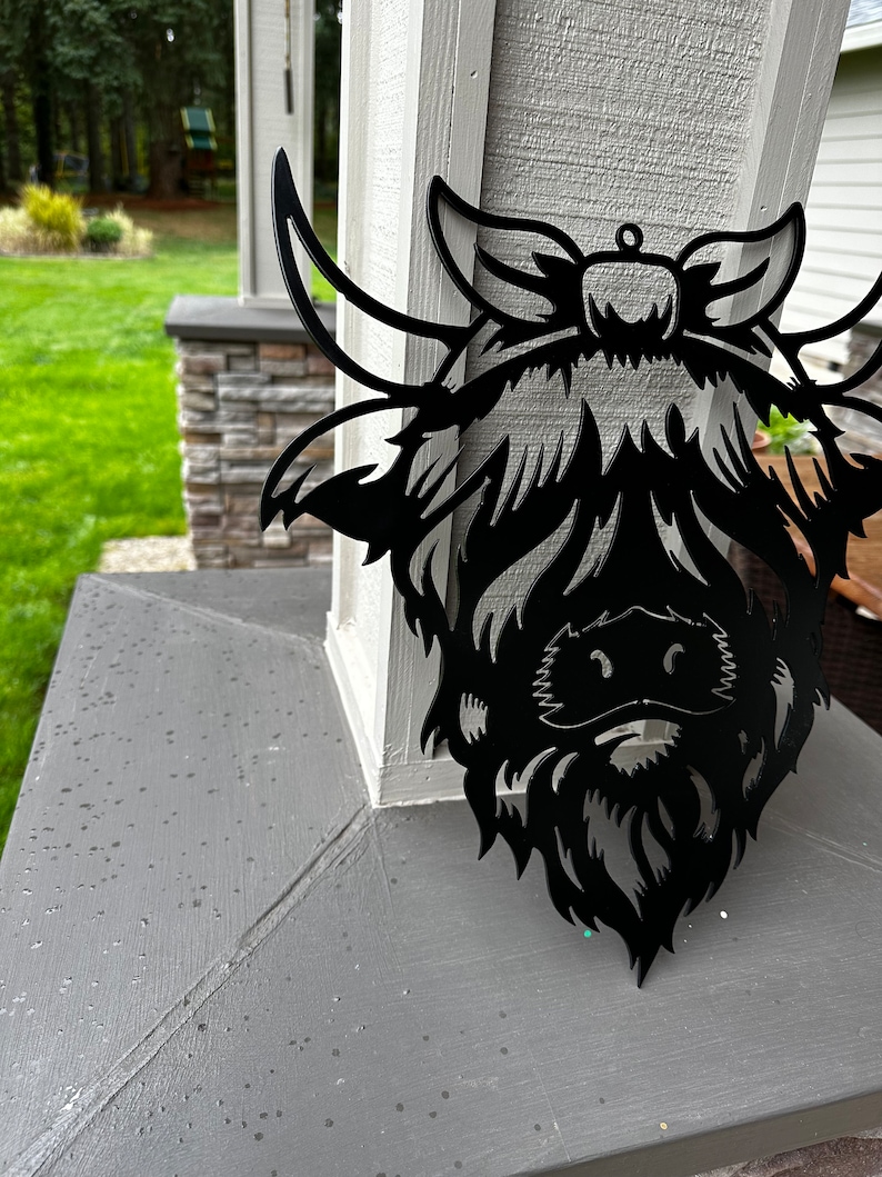 May include: Black metal silhouette of a Highland cow head with a bow on its head. The silhouette is mounted on a gray surface.