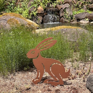 May include: A brown metal hare garden sculpture in a natural outdoor setting. The hare is in a seated position with long ears and detailed features. The sculpture is placed in a garden with greenery, rocks, and a small waterfall in the background.