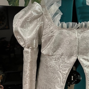 May include: A white brocade jacket with a ruffled collar and long sleeves. The jacket has a fitted bodice and a flowing skirt.