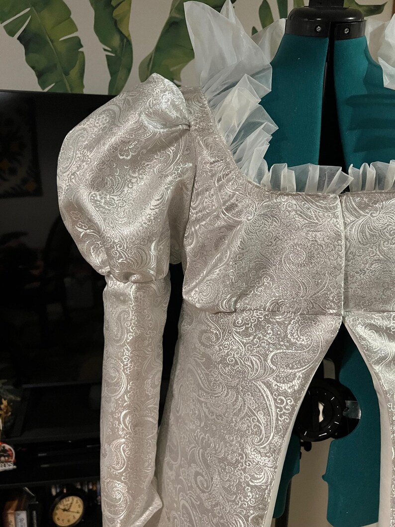 May include: A white brocade jacket with a ruffled collar and long sleeves. The jacket has a fitted bodice and a flowing skirt.