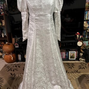 May include: A white gown with a high collar and long sleeves. The gown has a brocade pattern and a flowing skirt.