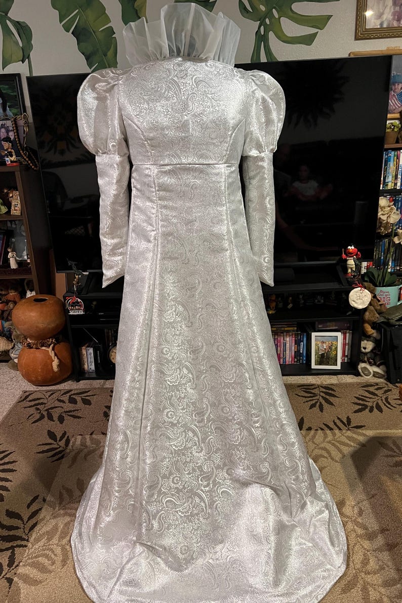 May include: A white gown with a high collar and long sleeves. The gown has a brocade pattern and a flowing skirt.