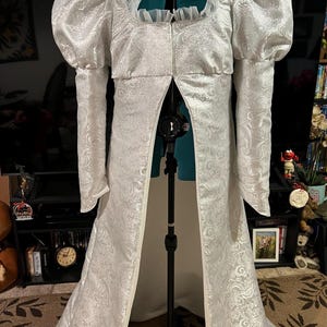 May include: A white, long-sleeved gown with a fitted bodice and a flowing skirt. The gown has a high neckline and a ruffled collar. The sleeves are puffed at the shoulders and the skirt is adorned with a paisley pattern.