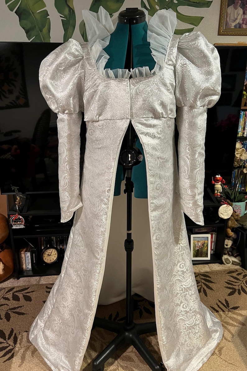 May include: A white, long-sleeved gown with a fitted bodice and a flowing skirt. The gown has a high neckline and a ruffled collar. The sleeves are puffed at the shoulders and the skirt is adorned with a paisley pattern.
