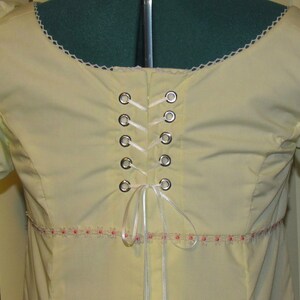 May include: A pale yellow blouse with a white lace trim around the neckline. The back of the blouse features a white ribbon laced through silver grommets.