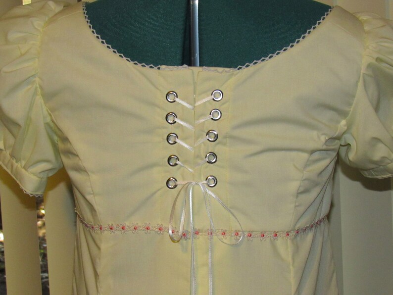 May include: A pale yellow blouse with a white lace trim around the neckline. The back of the blouse features a white ribbon laced through silver grommets.