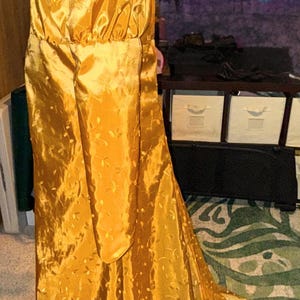 May include: A long, golden yellow gown with a gathered bodice and long sleeves. The gown is made of a shiny, satin-like fabric and has a delicate floral pattern.