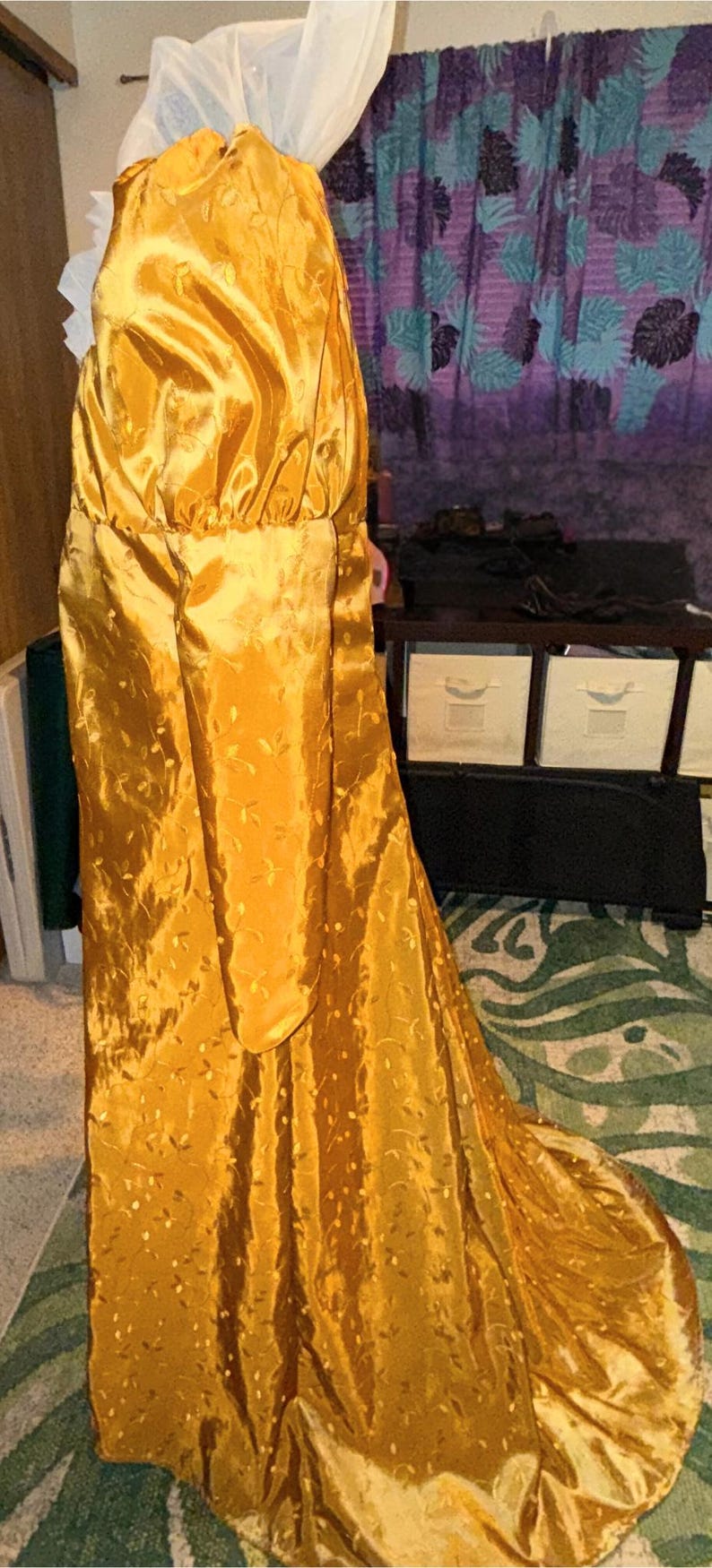 May include: A long, golden yellow gown with a gathered bodice and long sleeves. The gown is made of a shiny, satin-like fabric and has a delicate floral pattern.