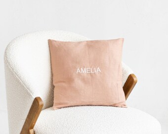 May include: A white armchair with a light pink pillow that says "Amelia" on it. The chair is on a white floor with a wicker basket and wooden blocks.