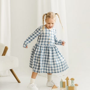 Può includere: Una bambina con un vestito a quadri blu e bianchi gioca con blocchi di legno. I blocchi sono impilati in una torre e sono dipinti in varie tonalità di verde, marrone e beige.