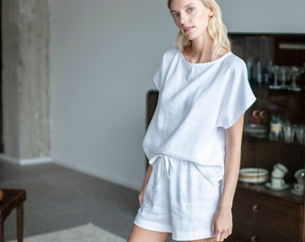 May include: A woman wearing a white linen top and matching shorts. She is standing in a room with a light grey carpet and a wooden cabinet with glass doors.