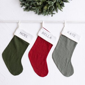 Pode incluir: Três meias de Natal penduradas em uma haste branca com uma guirlanda verde acima. As meias são verdes, vermelhas e cinzas com detalhes brancos e os nomes "Noel", "Bella" e "Kate" escritos nelas.