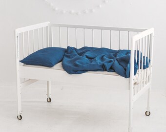 May include: A white cot with a blue linen bedding set. The cot has wheels and is on a white floor. There is a white rug with a blue toy piano on it.
