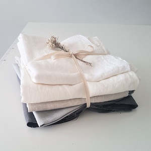 Puede incluir: Una pila de cuatro cuadrados de tela en varios tonos de gris y blanco, atados con un lazo de cuerda natural y una pequeña rama de flores secas.