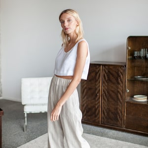 May include: A woman wearing a white linen tank top and beige linen trousers. She is standing in a room with a white wall and a wooden cabinet.