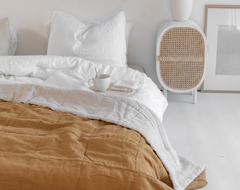 Könnte beinhalten: Ein weißes Bett mit einer braunen Bettwäsche aus Leinen, weißen Leinenlaken und einem weißen Kissen. Ein weißer Nachttisch mit einer geflochtenen Rattantür steht neben dem Bett. Eine weiße Vase mit getrockneten Blumen steht auf dem Nachttisch.