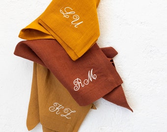 May include: Three linen napkins in mustard yellow and rust brown colors, folded and stacked. The napkins are embroidered with the initials "LOU", "RM", and "KL" in white thread.