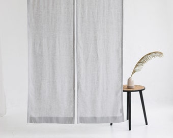 Puede incluir: Dos cortinas de lino gris claro con ribete gris oscuro colgadas de una barra blanca. Las cortinas están delante de una pared blanca y una pequeña mesa auxiliar negra con un jarrón y una rama seca.