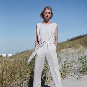 May include: A woman wearing a light gray linen jumpsuit with a wide belt and black sandals. She is standing on a sandy beach with tall grass in the background.