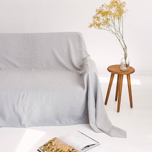 May include: A light gray linen sofa throw draped over a couch with a small wooden stool and a vase of yellow flowers in the background.