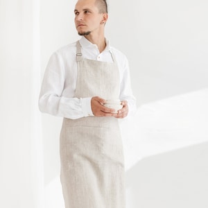 Pode incluir: Uma pessoa vestindo uma camisa branca de linho e um avental de linho bege. O avental tem um bolso grande na frente e alças ajustáveis. A pessoa está segurando uma tigela branca pequena em suas mãos.