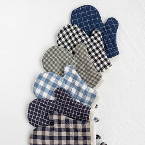 Può includere: Una collezione di guanti da forno trapuntati con vari motivi a quadri, tra cui bianco e nero, blu e beige. I guanti sono disposti in una fila diagonale su uno sfondo bianco, mostrando i loro diversi design e colori.