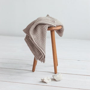 May include: A light brown waffle weave throw draped over a wooden stool with three legs. The stool is sitting on a white wooden floor.