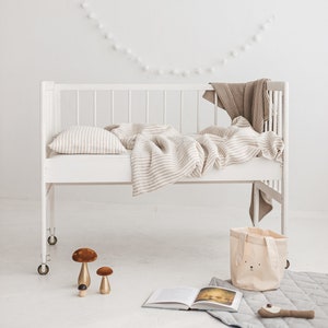 May include: A white crib with a beige and white striped linen bedding set. The crib is decorated with a string of white pom-poms and a framed picture of a dog. There is a beige canvas tote bag with a bunny face on it and a book on the floor.