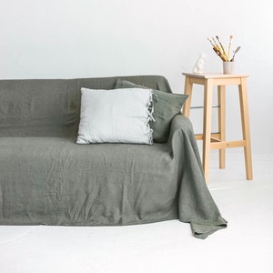 May include: A green linen sofa with a white pillow and a wooden stool with a white ceramic figurine and paintbrushes in a glass jar on top.