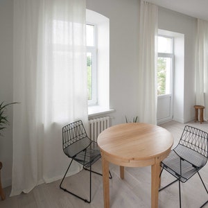 May include: A round wooden table with two black wire chairs sits in a white room with large windows and white curtains. The floor is light wood.