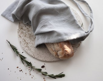Pode incluir: Uma bolsa de pão de linho cinza com fecho de cordão, contendo um pão sobre um prato de cerâmica branca salpicado. Um ramo de alecrim e migalhas de pão espalhadas sobre a superfície branca.