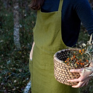 May include: A person wearing a green linen apron with a large pocket on the left side, holding a woven basket filled with orange berries and green leaves.