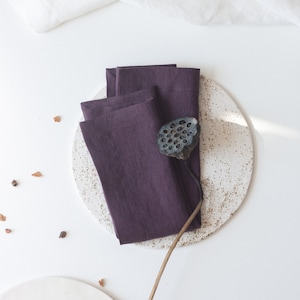 May include: A set of three purple linen napkins folded on a white speckled plate with a dried lotus pod on top.