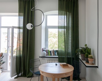 May include: A round wooden dining table with two black metal chairs. The table is set with a book and a small object. The room has green curtains, a window with a view of trees, and a shelf with books.