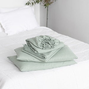Peut inclure: Un lit blanc avec un ensemble de literie en lin vert sauge. La literie est pliée soigneusement sur le lit. Le lit est dans une pièce blanche avec une petite plante dans le coin.