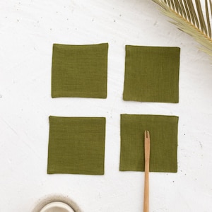 Puede incluir: Cuatro posavasos cuadrados de lino verde oliva con un utensilio de madera con un tenedor en el extremo.