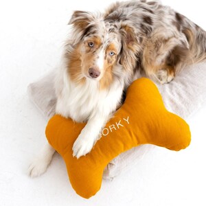 Könnte beinhalten: Ein Australian Shepherd mit blauem Merle liegt auf einem beigefarbenen Kissen mit einem gelben knochenförmigen Kissen, auf dem das Wort "WORKY" gestickt ist.