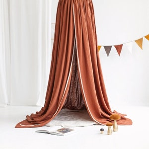 Puede incluir: Una tienda de campaña de lona de color terracota con ribete blanco, colgada del techo. La tienda está drapeada sobre una alfombra blanca con un libro y setas decorativas en el suelo.