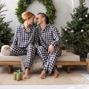 May include: A couple wearing matching navy and white plaid pajamas sit on a wooden bench. A green wreath hangs on the wall behind them, with a small Christmas tree to the right. A wrapped gift sits on the rug in front of them.