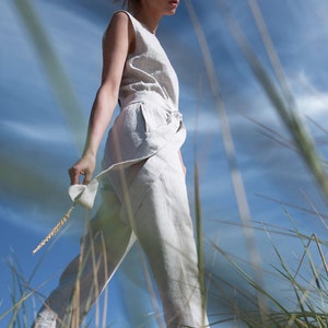 May include: A woman wearing a white linen jumpsuit walks through tall grass with a blue sky in the background.