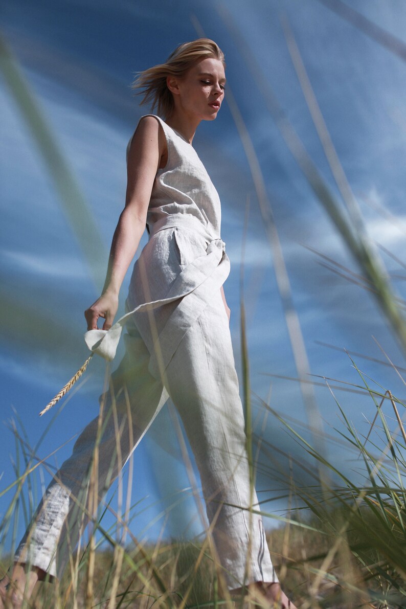 May include: A woman wearing a white linen jumpsuit walks through tall grass with a blue sky in the background.