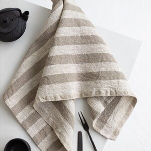 May include: A beige linen tea towel with white stripes folded on a white surface with black cutlery and a black tea pot.