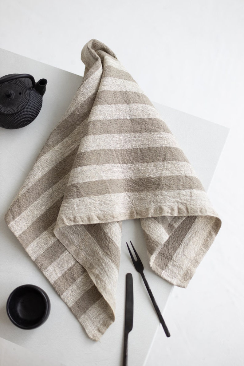 May include: A beige linen tea towel with white stripes folded on a white surface with black cutlery and a black tea pot.