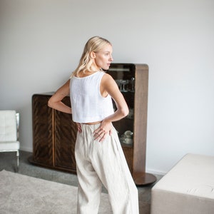 K&ouml;nnte beinhalten: Ein wei&szlig;es Leinen-Crop-Top und eine beige Leinenhose. Das Outfit wird von einer Person getragen, die in einem Raum mit einem Holzschrank und einem neutralfarbenen Teppich steht. Die Hose hat ein weites Bein-Design.