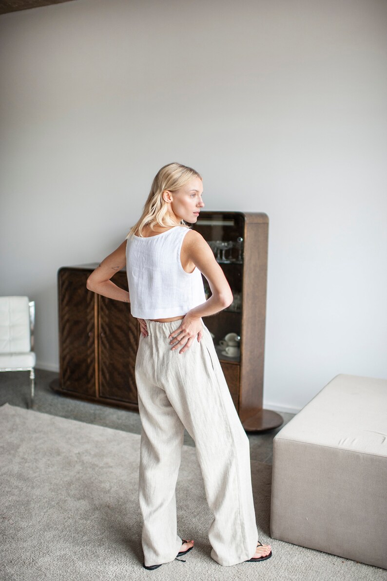 K&ouml;nnte beinhalten: Ein wei&szlig;es Leinen-Crop-Top und eine beige Leinenhose. Das Outfit wird von einer Person getragen, die in einem Raum mit einem Holzschrank und einem neutralfarbenen Teppich steht. Die Hose hat ein weites Bein-Design.