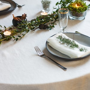 May include: A white tablecloth with a simple place setting, featuring a white napkin, a fork, and a black plate. A sprig of greenery decorates the napkin. A garland of greenery with small candles and a glass of water is on the table.