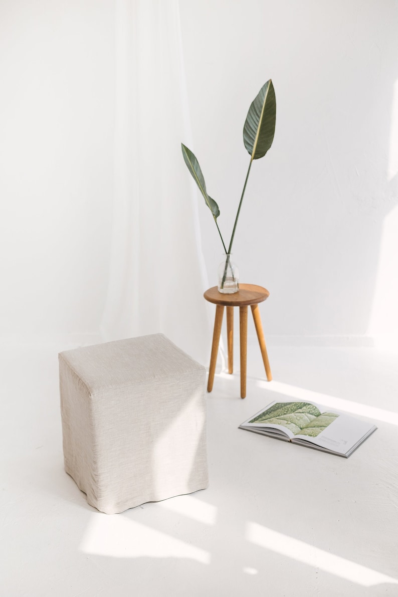 May include: A white linen ottoman sits on a white floor with a small wooden stool and a vase with green leaves. A book with a green leaf design is on the floor.